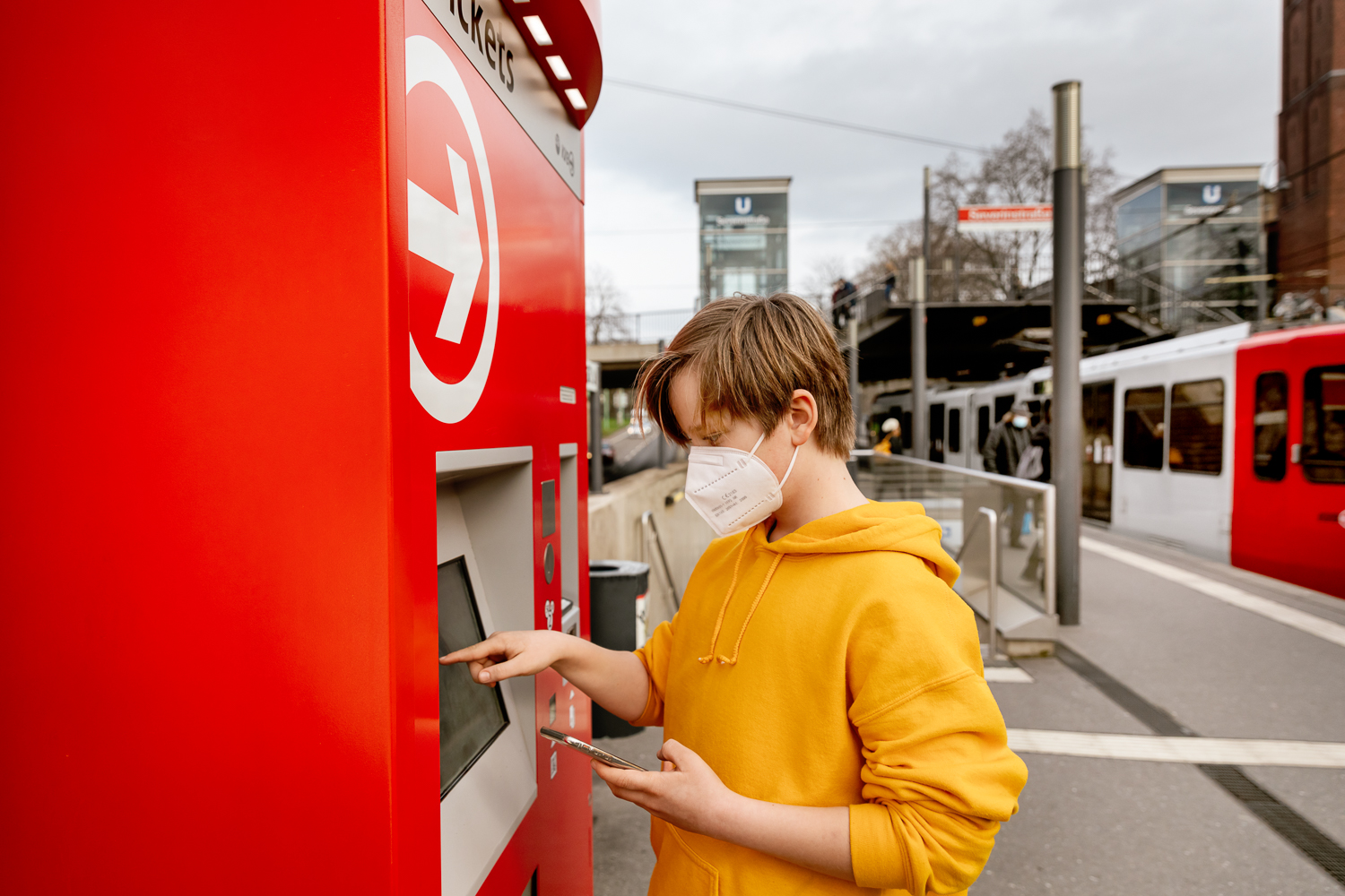 Wir sehen einen jugendlichen Fahrgast mit Mundschutz, wie er auf einem leeren Bahnsteig ein Ticket zieht.