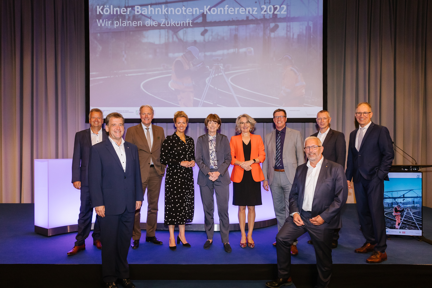 Ein Gruppenbild auf dem Podium zeigt die prominenten Gäste: Heiko Sedlaczek, Geschäftsführer Nahverkehr Rheinland, Bernd Köppel, Leiter Infrastrukturprojekte West DB Netz AG, Gerald Böse, Vorsitzender der Geschäftsführung Koelnmesse GmbH, Ina Brandes, damalige Ministerin für Verkehr des Landes Nordrhein-Westfalen, Henriette Reker, Oberbürgermeisterin der Stadt Köln, Susanne Henckel, Staatssekretärin im Bundesministerium für Digitales und Verkehr, Stephan Santelmann, Verbandsvorsteher Zweckverband Nahverkehr Rheinland, Manfred Gutfrucht, Leiter Portfolio Köln der DB Netz AG, Frank Sennhenn, Vorstandsvorsitzender DB Netz AG, Dr. Norbert Reinkober, Geschäftsführer Nahverkehr Rheinland